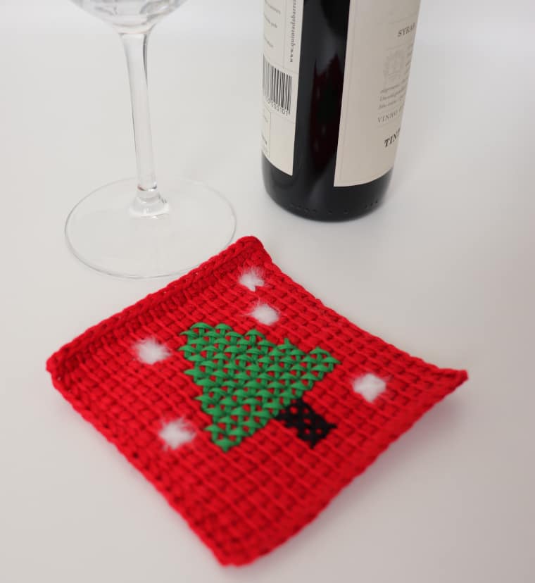 The Tunisian Pine Tree Christmas Coaster on a white table and next to a glass of wine and a small bottle of wine