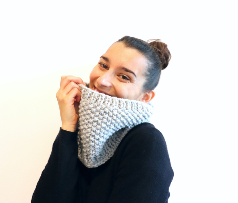 Susana of Fluffy Stitches wearing the Knit Easy Seed Cowl in light grey and a black sweater against a white background.
