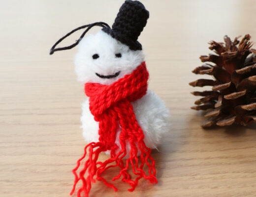 A smiling Crochet Snowman Christmas Ornament, made of white faux fur with a striking red bulky scarf and a black top hat next to a pine cone.