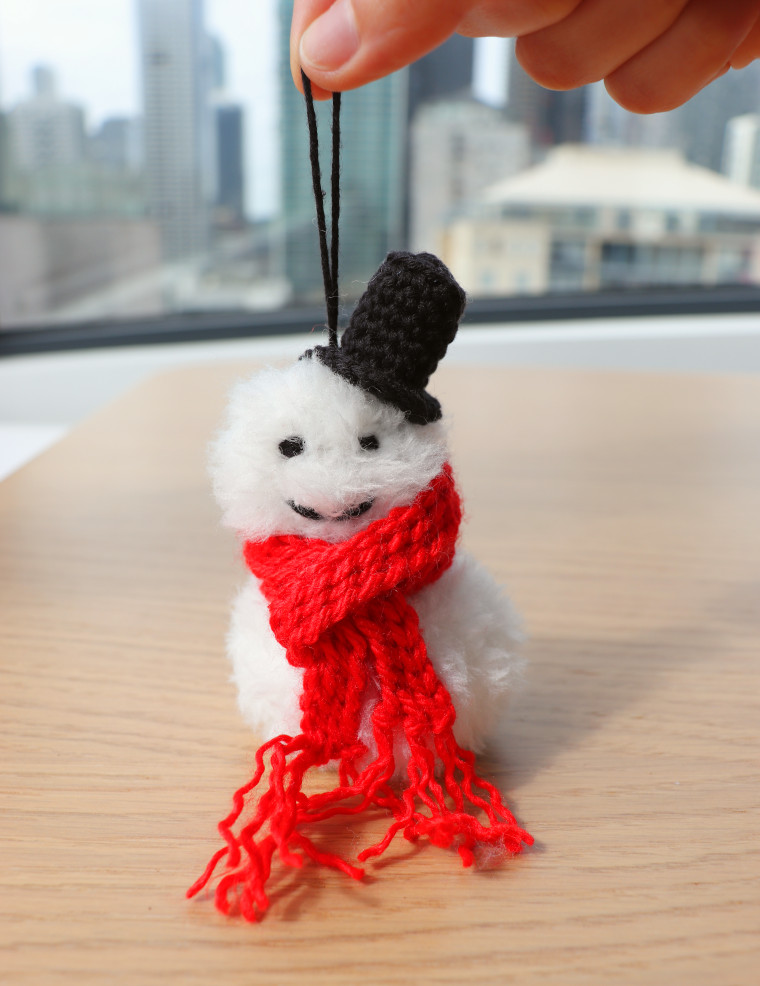 A smiling Crochet Snowman Christmas Ornament, made of white faux fur with a striking red bulky scarf and a black top hat.